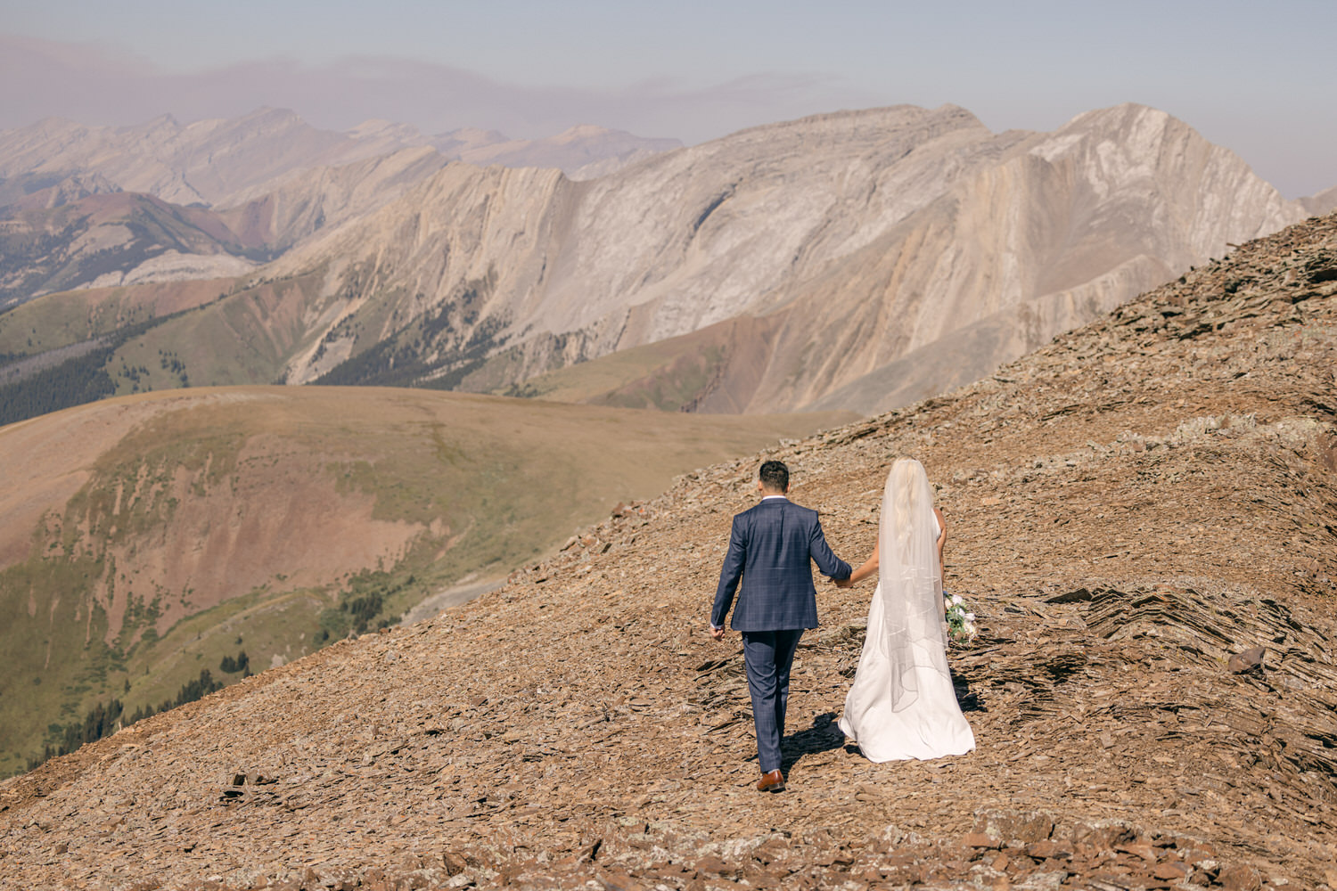 A couple walking hand in hand on a rocky mountain ridge, surrounded by breathtaking peaks and valleys.