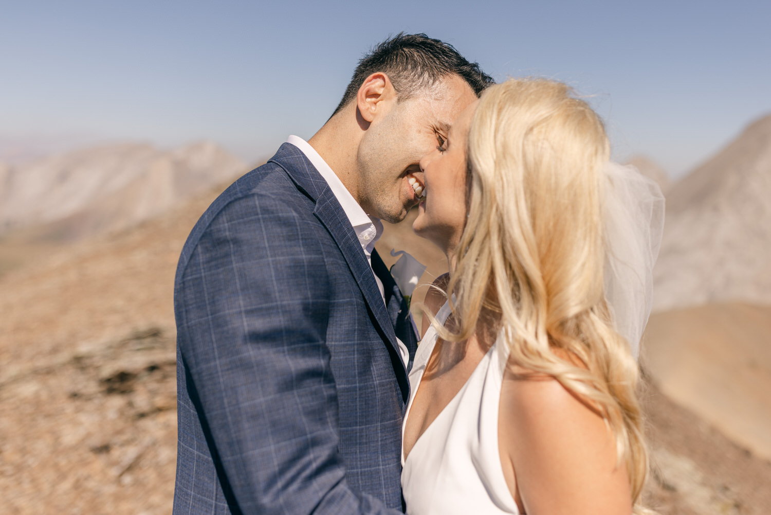 A bride and groom embrace and share a kiss in a serene mountain setting, showcasing love and connection against a backdrop of majestic peaks.