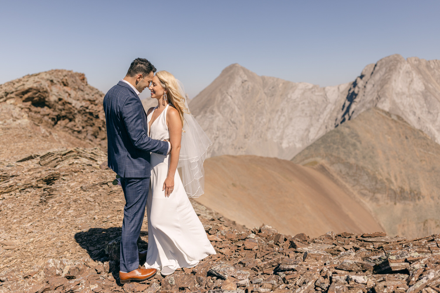 A couple stands lovingly together in wedding attire on a rocky mountain peak, with majestic peaks in the background under a clear blue sky.