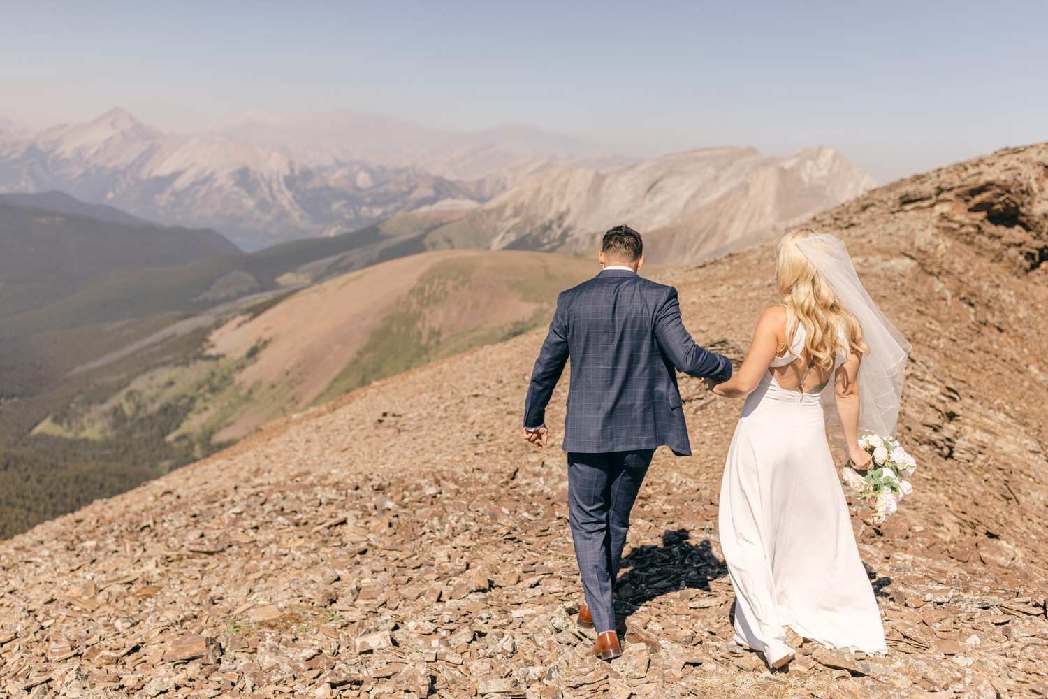 A couple dressed elegantly, walking hand in hand along a rocky mountain ridge with breathtaking landscapes in the background.