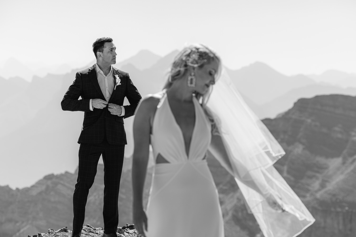 A groom adjusts his suit while his bride, dressed in a sleek gown and veil, turns away, set against a stunning mountain backdrop.