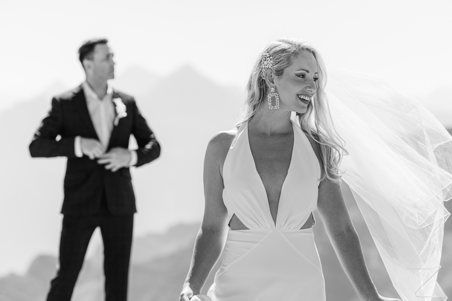 A smiling bride in a flowing white dress and veil stands in a scenic outdoor setting, with a groom adjusting his jacket in the background.