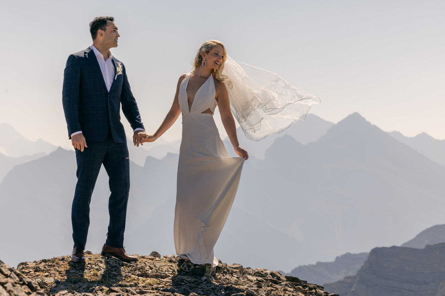 A couple holding hands on a rocky mountain summit, with the bride's veil flowing in the wind amid a scenic mountainous backdrop.