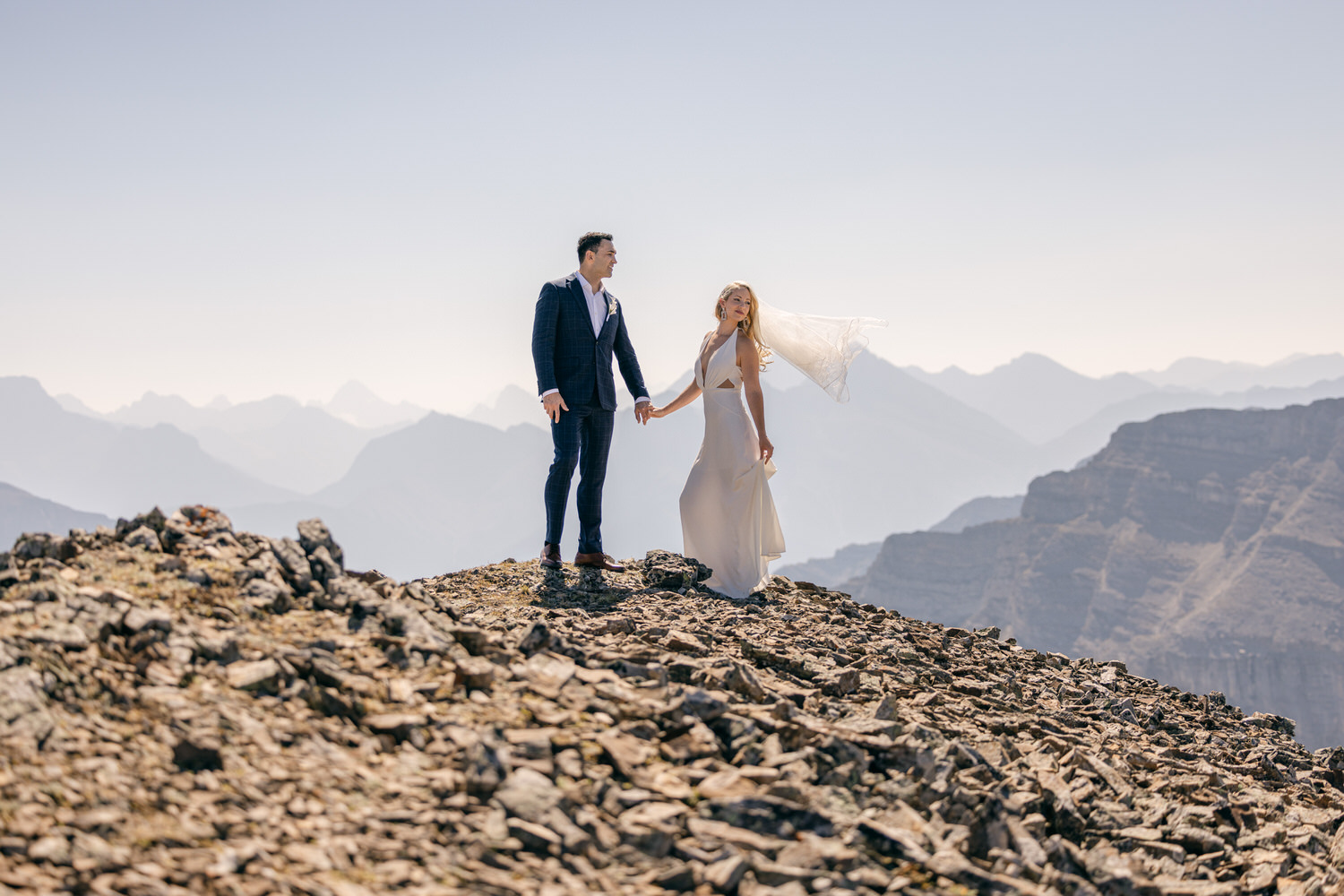 A couple stands hand-in-hand on a rocky mountain peak, surrounded by breathtaking scenery and a soft breeze flowing through the bride's veil.