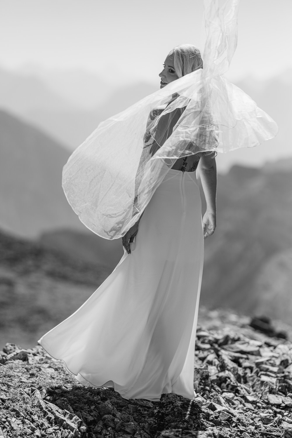 A woman in a flowing white dress poses atop rocky mountains, with a sheer veil billowing in the wind, against a soft-focus backdrop of distant peaks.