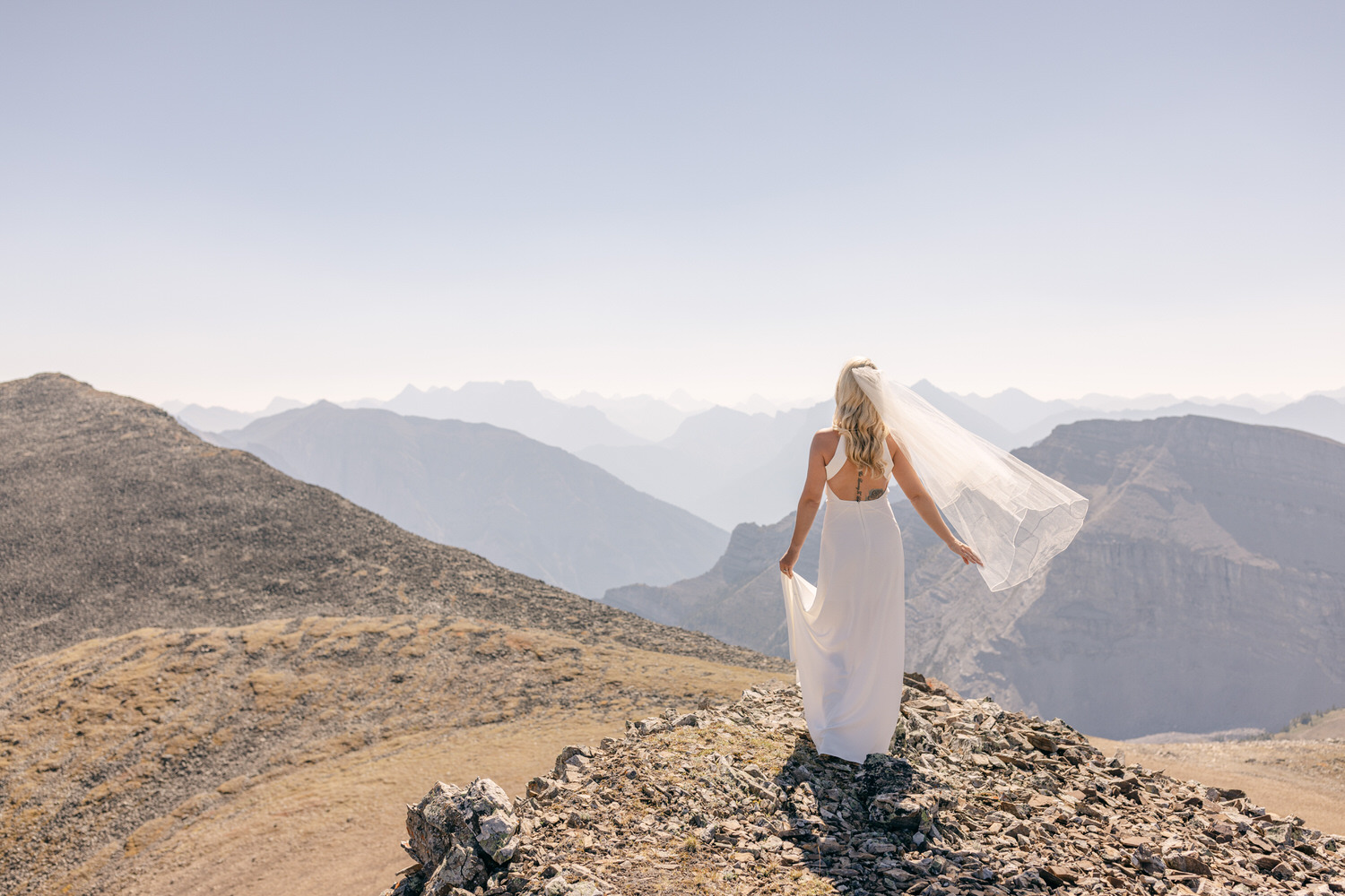 A bride in a flowing white dress and veil stands on a rocky mountain peak, gazing at the expansive landscape of distant mountains under a clear sky.