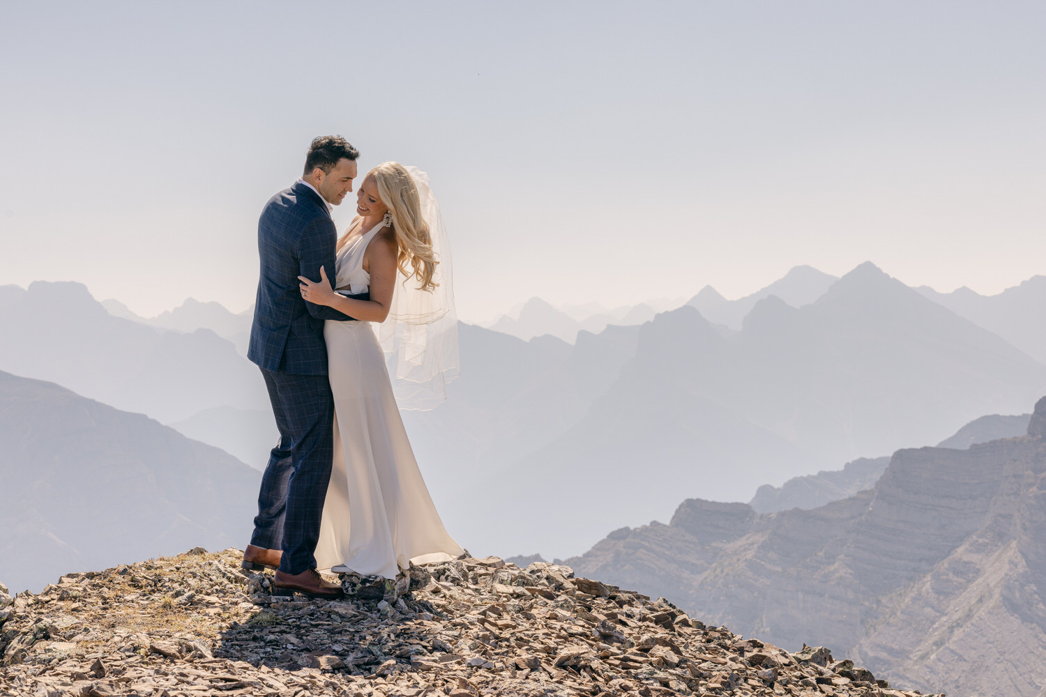 A bride and groom embrace on a rocky mountain peak, surrounded by a breathtaking landscape of distant peaks and soft haze.