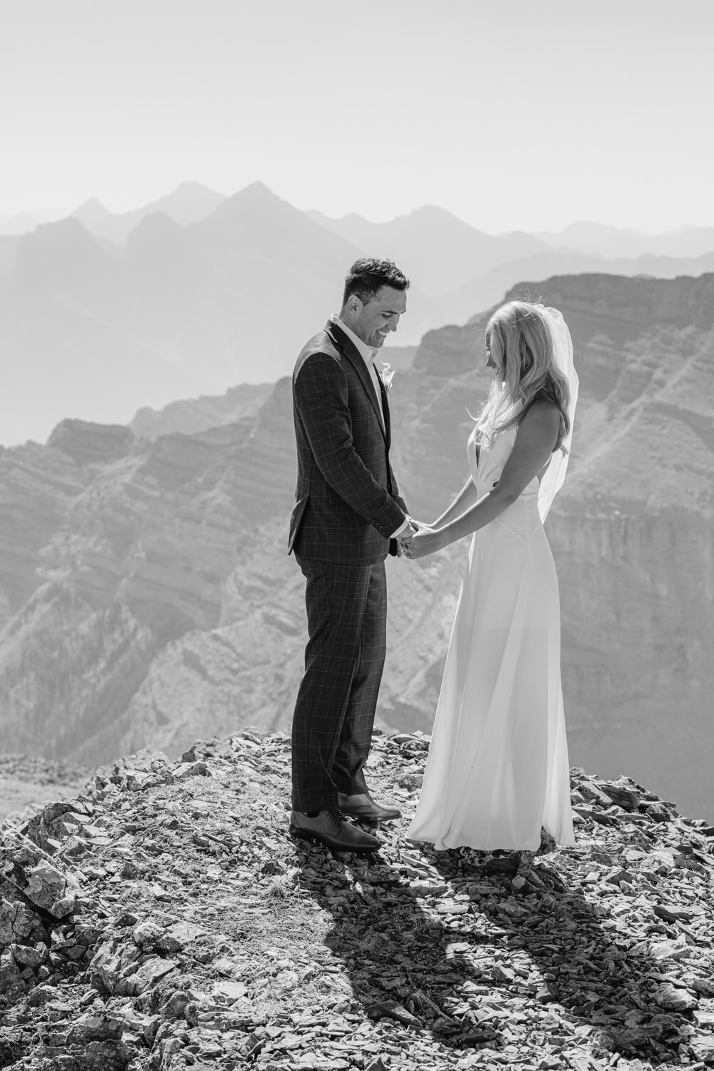 A couple holds hands on a rocky mountain ledge, exchanging vows with a breathtaking mountain range in the background, captured in black and white.