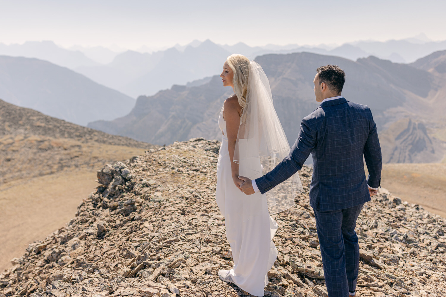 A bride and groom holding hands on a rocky mountain ridge, with breathtaking mountain scenery in the background.