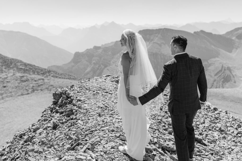 A bride and groom holding hands on a rocky mountain peak, with stunning mountain vistas in the background.