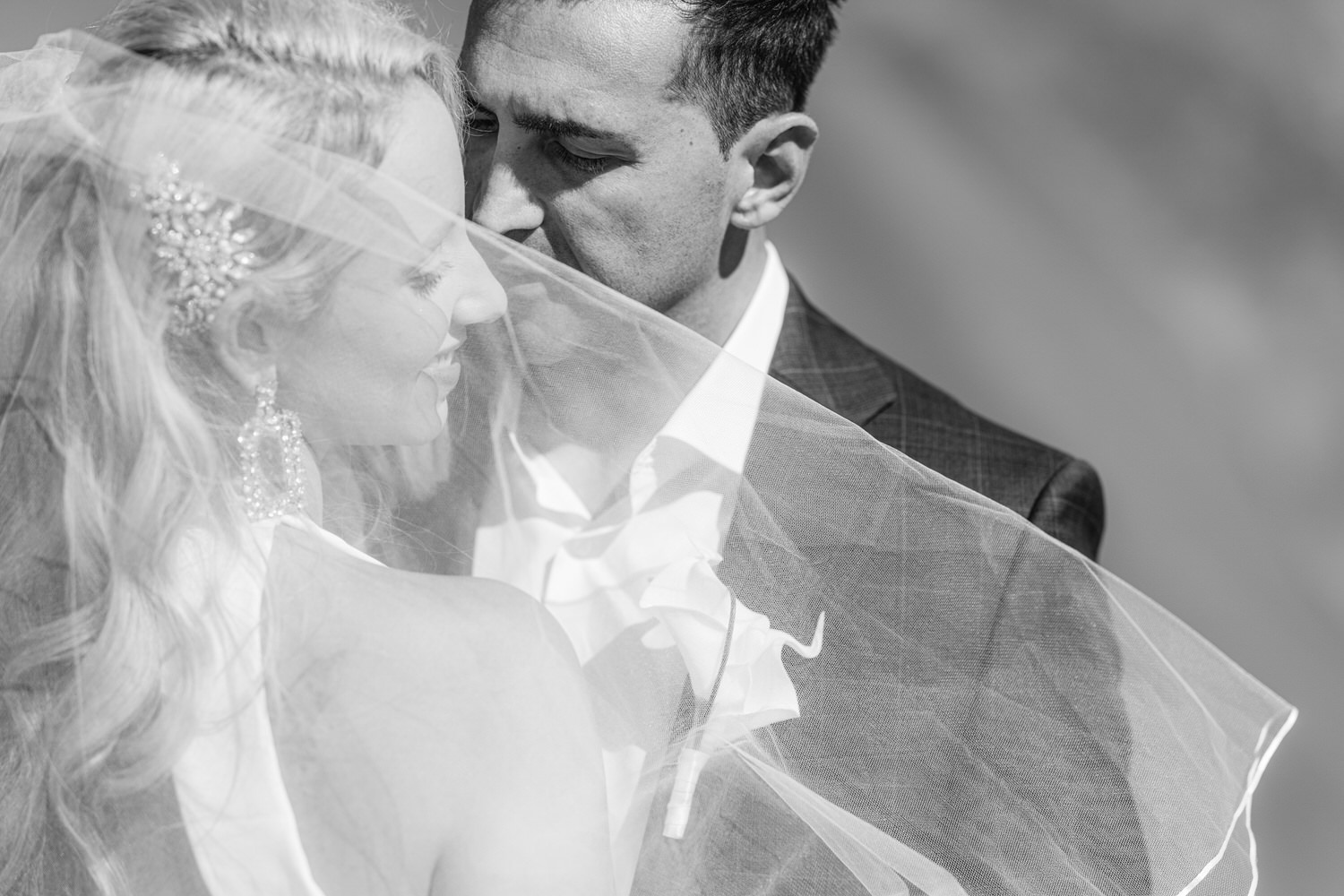 A couple sharing a tender embrace, partially obscured by a delicate wedding veil, capturing the essence of love and connection in a monochrome setting.