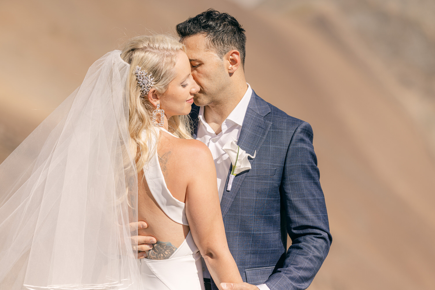 A bride and groom share an intimate moment, with the bride in a stunning gown and veil, while the groom wears a blue plaid suit, set against a beautiful natural backdrop.