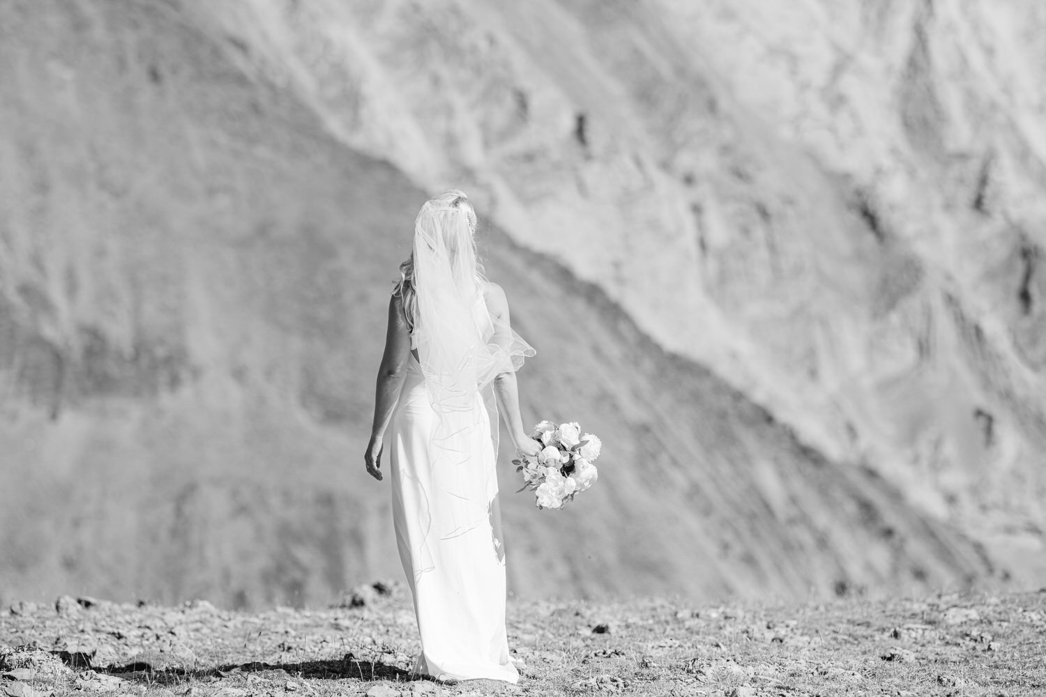 A bride in a flowing white gown and veil stands with her back to the camera, holding a bouquet of flowers against a dramatic mountainous backdrop.