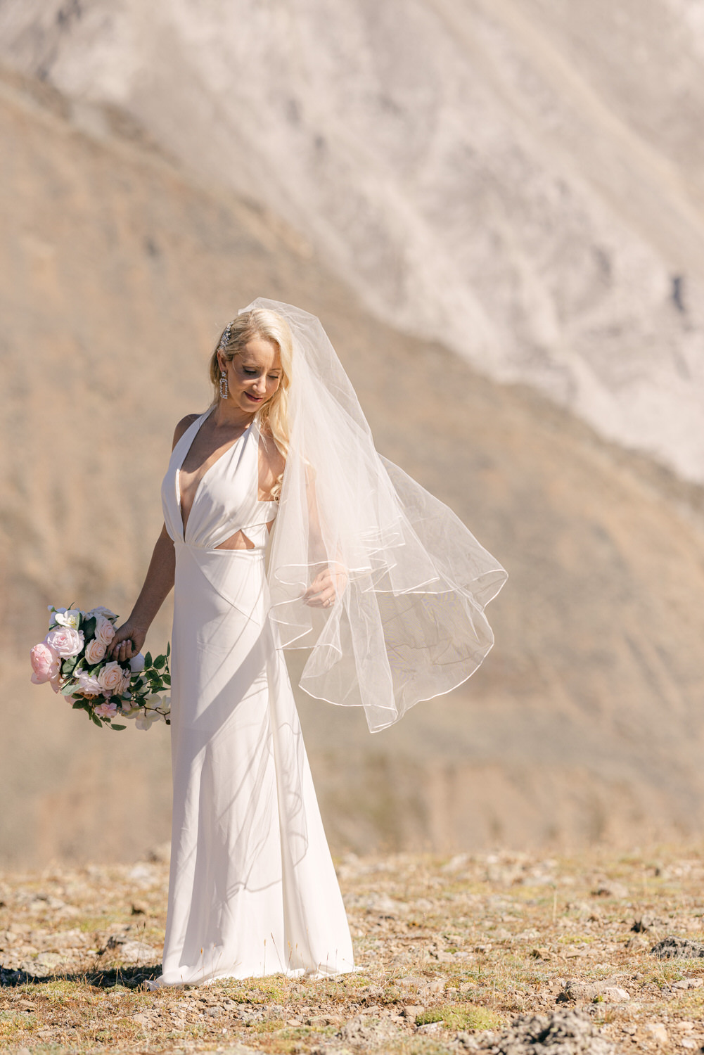 A bride in a flowing white dress and veil holds a bouquet of pink roses, standing gracefully on rocky terrain with mountains in the background.