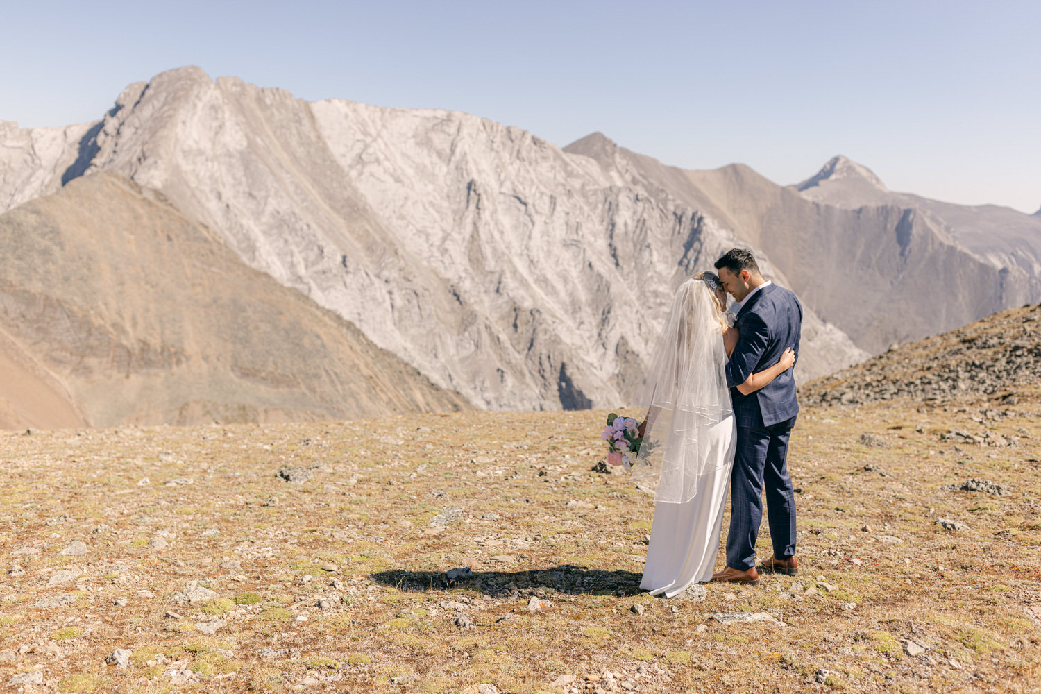 A couple shares a romantic embrace on a mountaintop, surrounded by stunning rocky peaks and a clear blue sky.