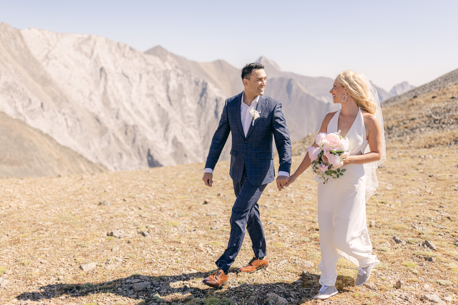 A couple joyfully walking hand in hand through a mountain landscape, with the bride in a white dress and bouquet, and the groom in a blue suit.