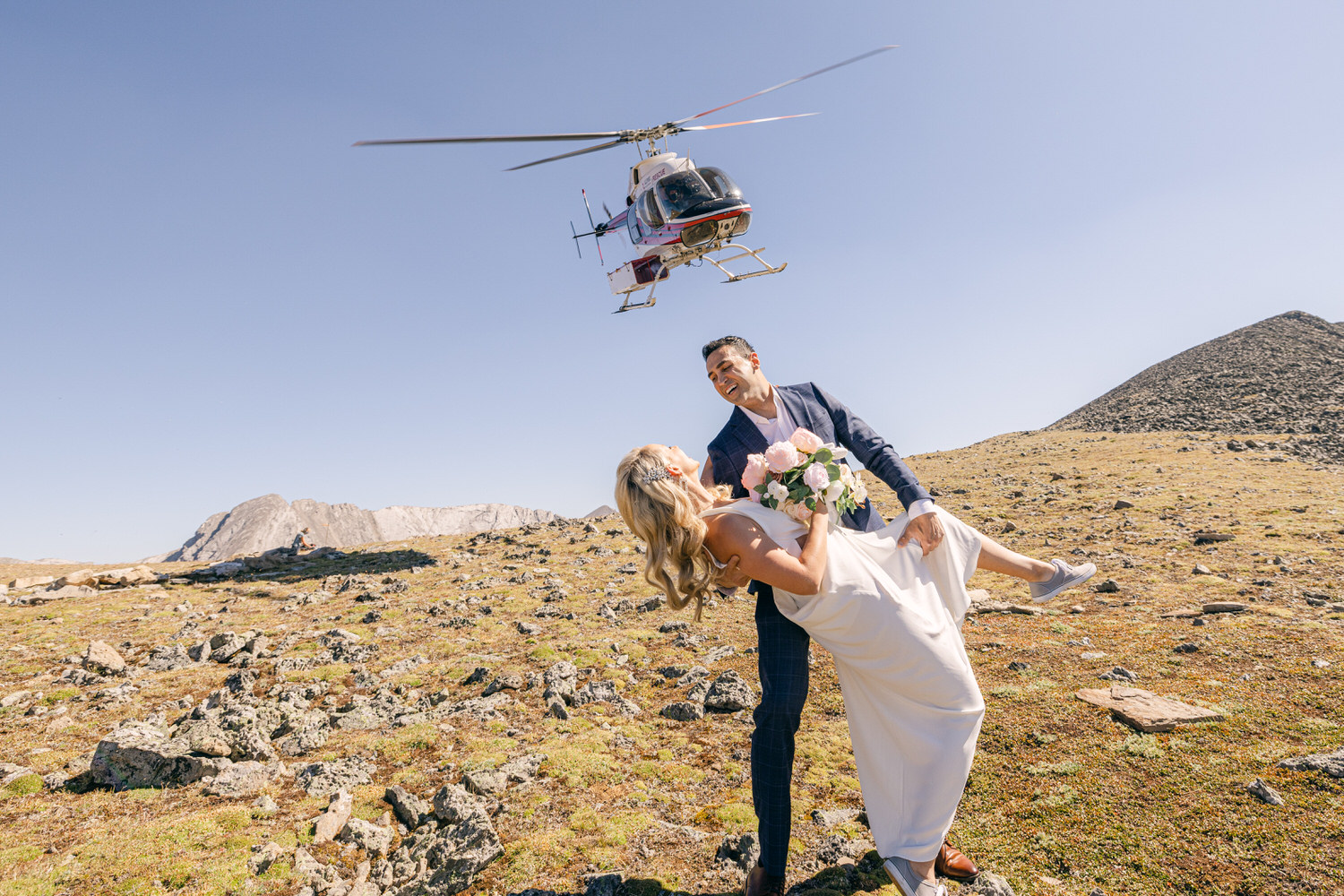 Mountain Wedding Adventure::A joyful couple embraces under a helicopter in a scenic mountain landscape, celebrating their wedding day.