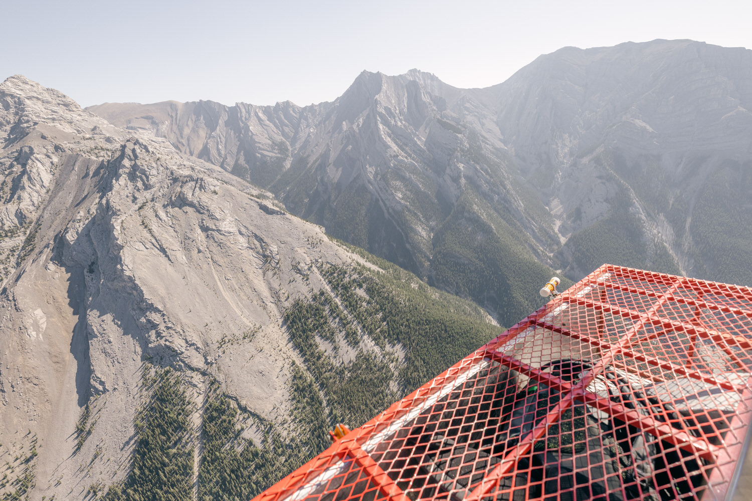 A panoramic view of rugged mountains from an elevated platform, featuring a red mesh surface in the foreground and lush trees stretching across the valley below.