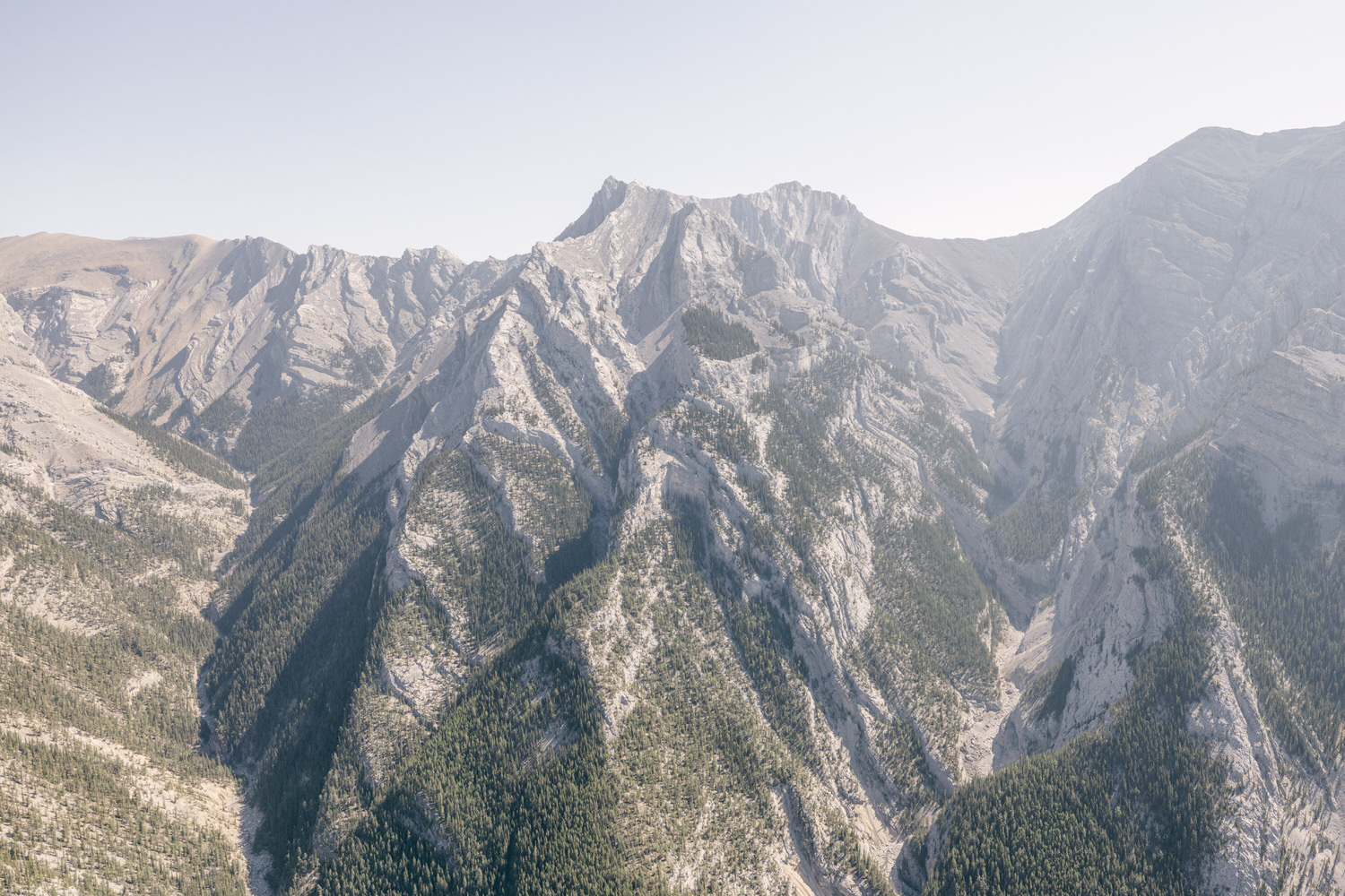 Aerial view of rugged mountains with sharp peaks and a lush green forest in the valleys, under a clear sky.