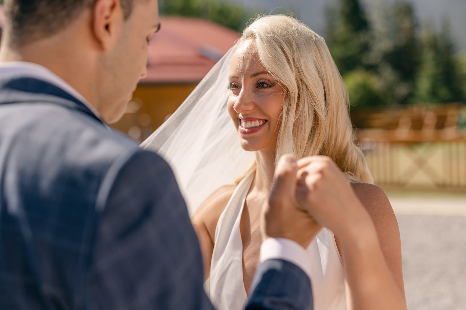 Bride and groom share a joyful moment outdoors, with the bride smiling radiantly while holding the groom's hands, surrounded by nature.