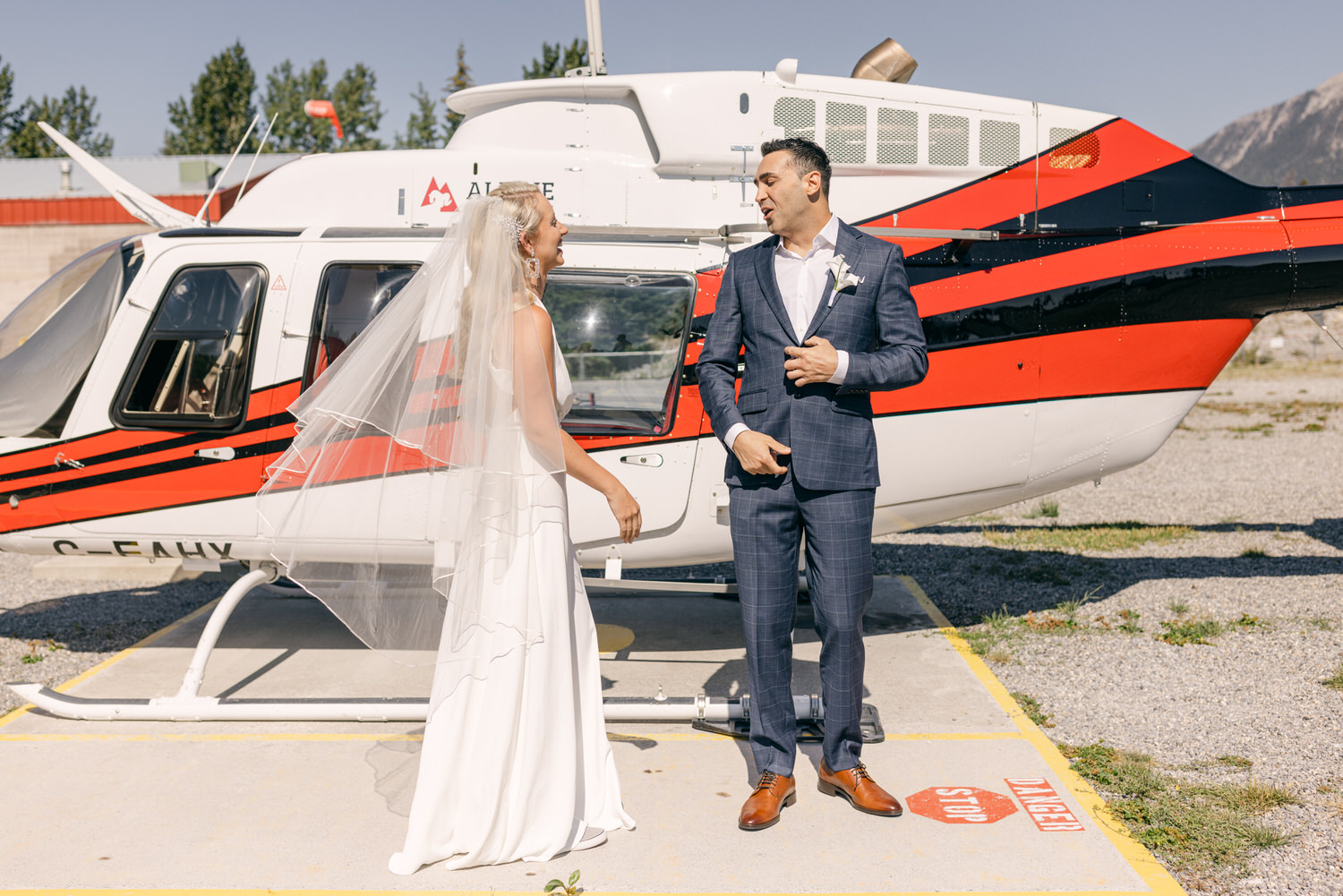 A bride in a flowing white dress and veil smiles at a groom in a blue checkered suit, standing next to a red and white helicopter in a sunny outdoor setting.