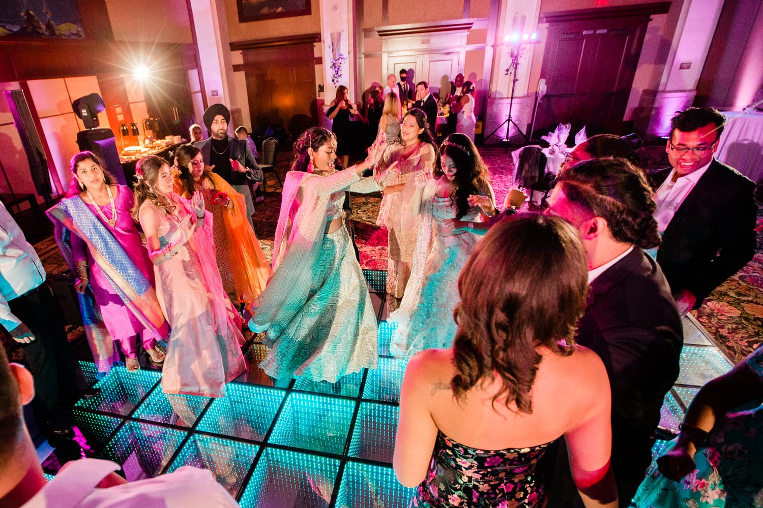 Guests in vibrant outfits enjoy a lively dance at a glamorous event, surrounded by colorful lights and an energetic atmosphere.