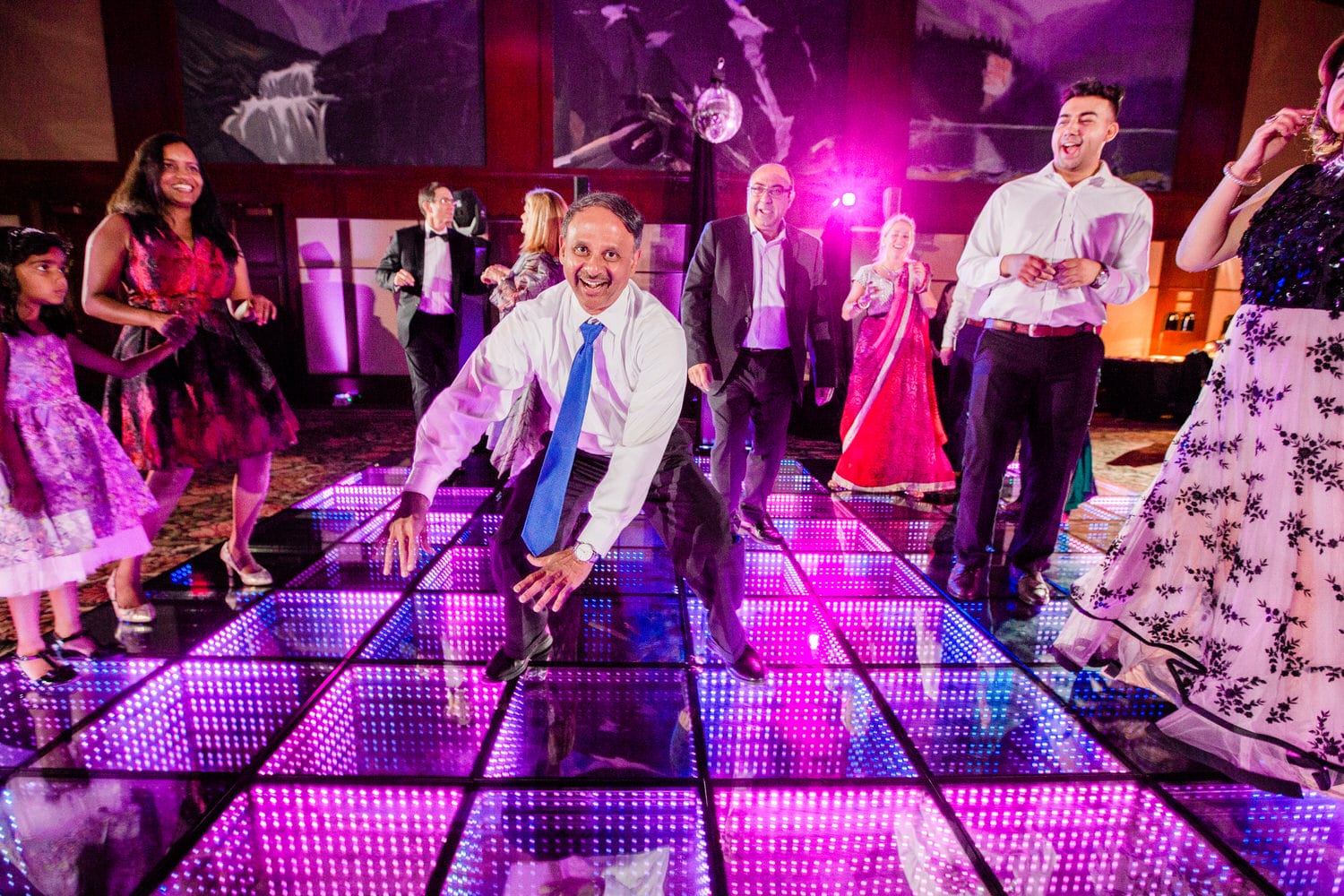 A lively scene of people enjoying a dance party on a colorful light-up dance floor, featuring both adults and children in festive attire.