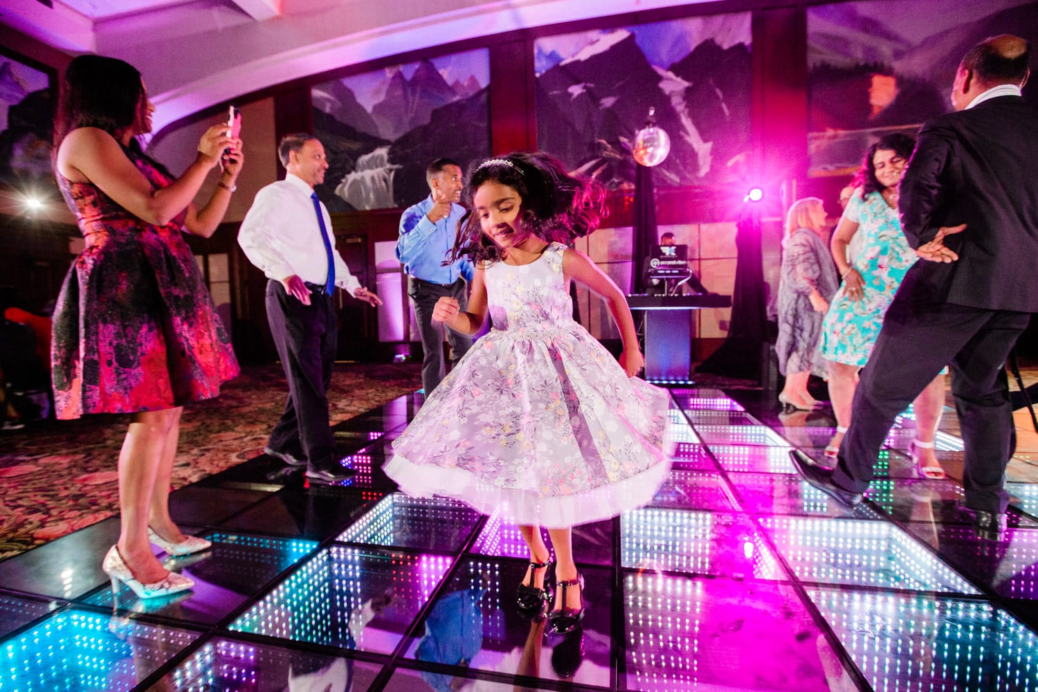 A lively dance scene featuring a young girl in a floral dress twirling while adults enjoy the celebration around her, illuminated by colorful lights.