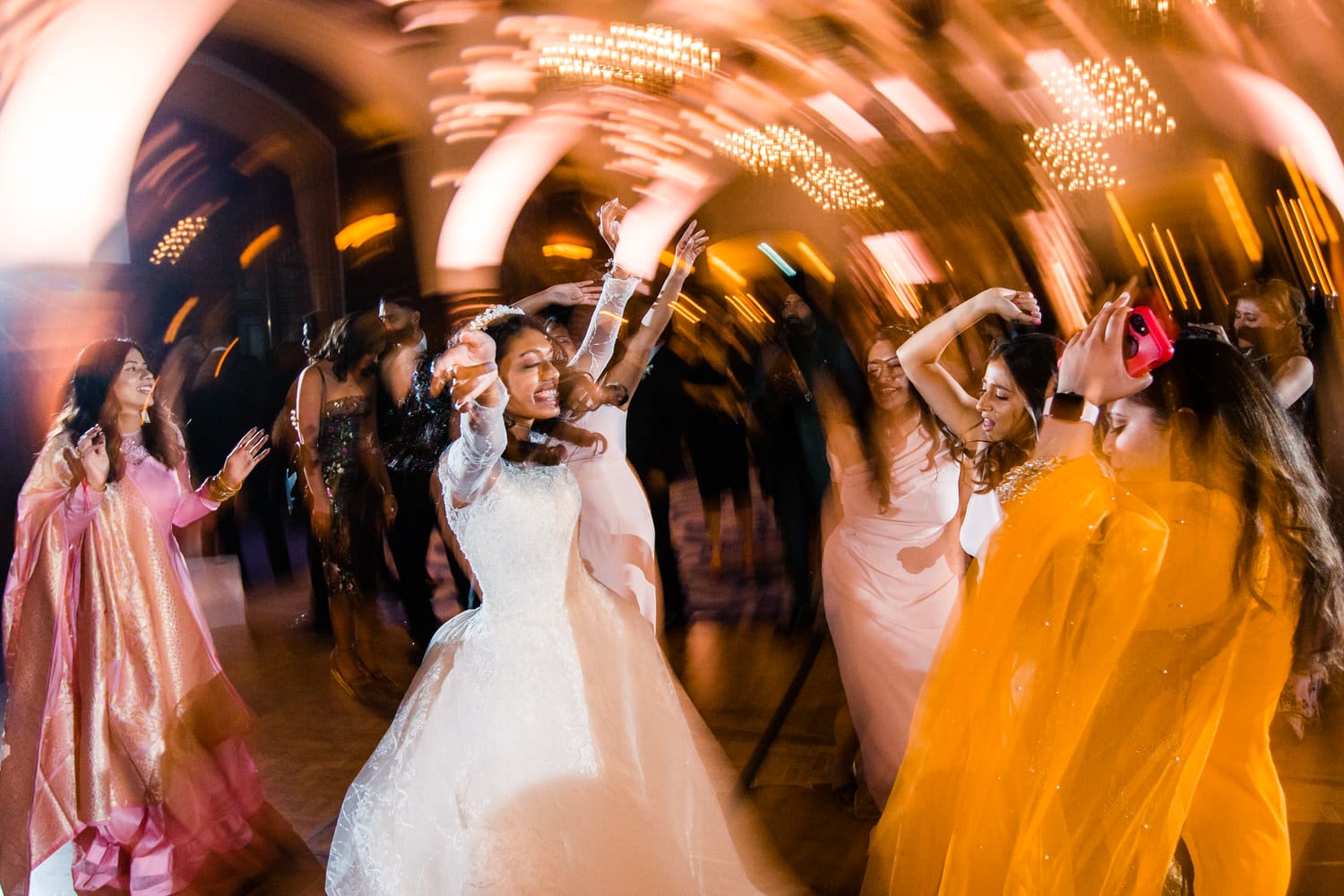 A bride and her friends dance energetically at a wedding reception, captured in a vibrant, motion-filled photograph.