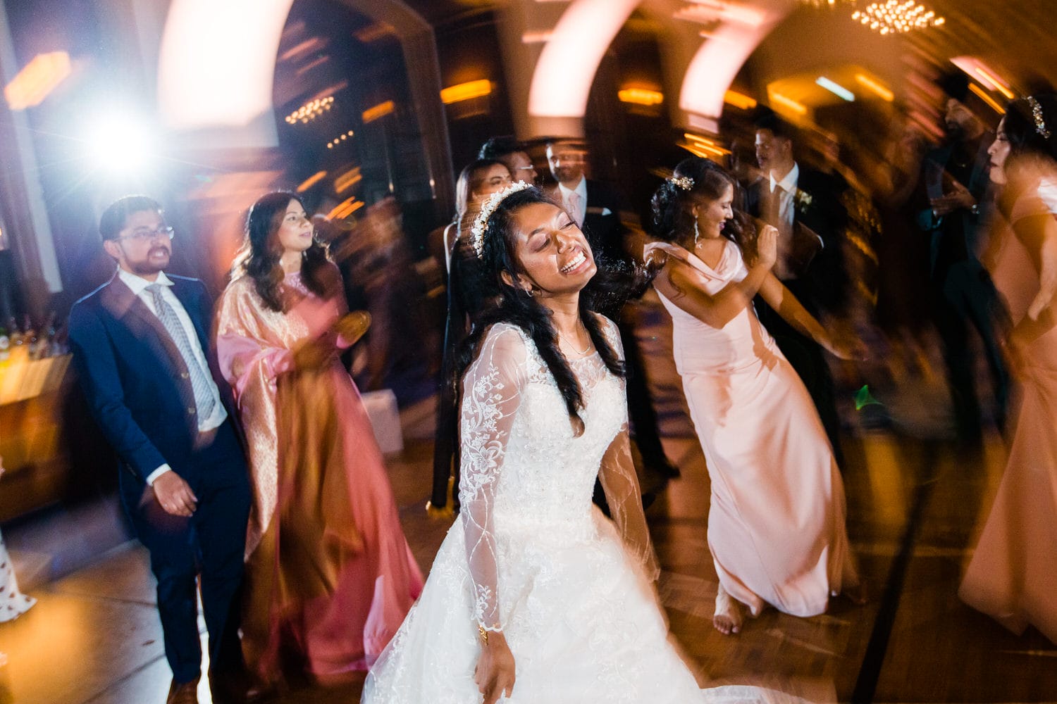 A bride dances joyfully among friends at a vibrant wedding reception, surrounded by colorful lights and festive ambiance.