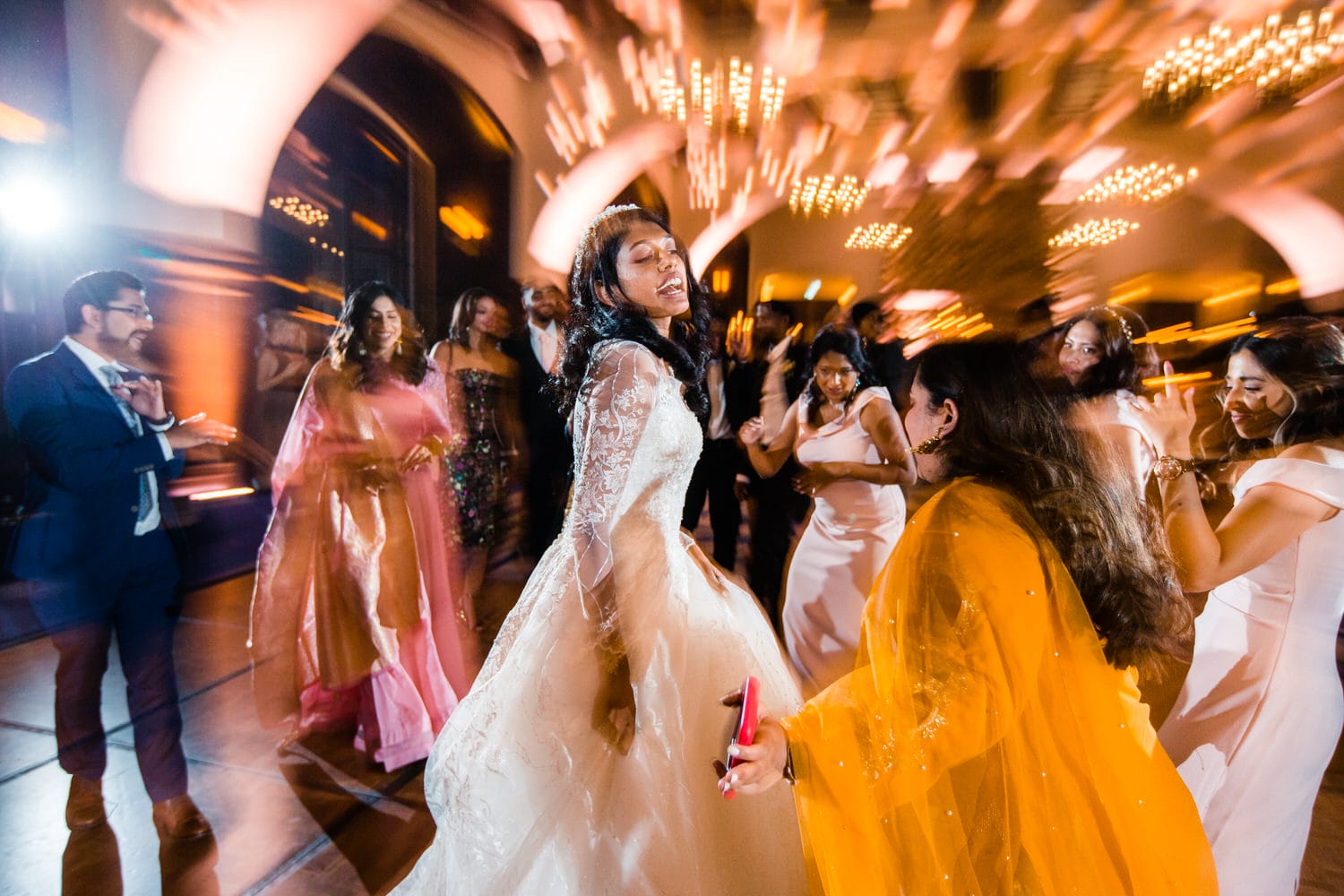 A bride dances joyfully among guests in a vibrant wedding reception, with swirling lights and festive attire creating a lively atmosphere.