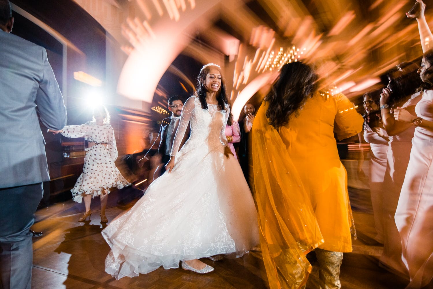 A joyful bride in a stunning white gown dances with friends, surrounded by motion blur and vibrant lights in a lively celebration.