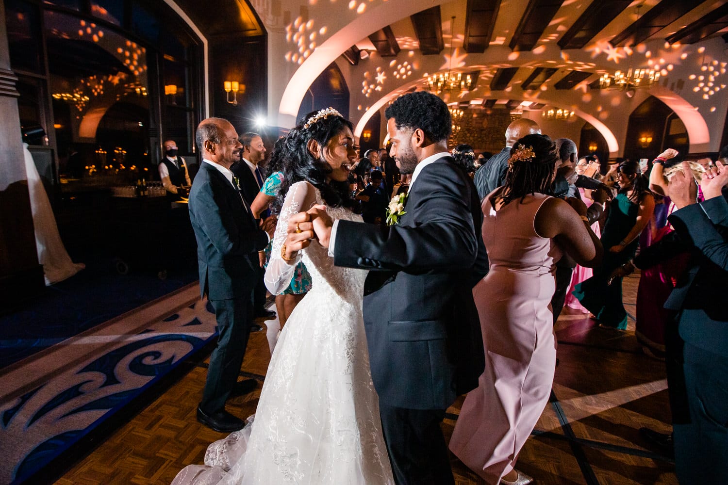 A bride and groom share a romantic dance surrounded by guests celebrating at a festive wedding reception.