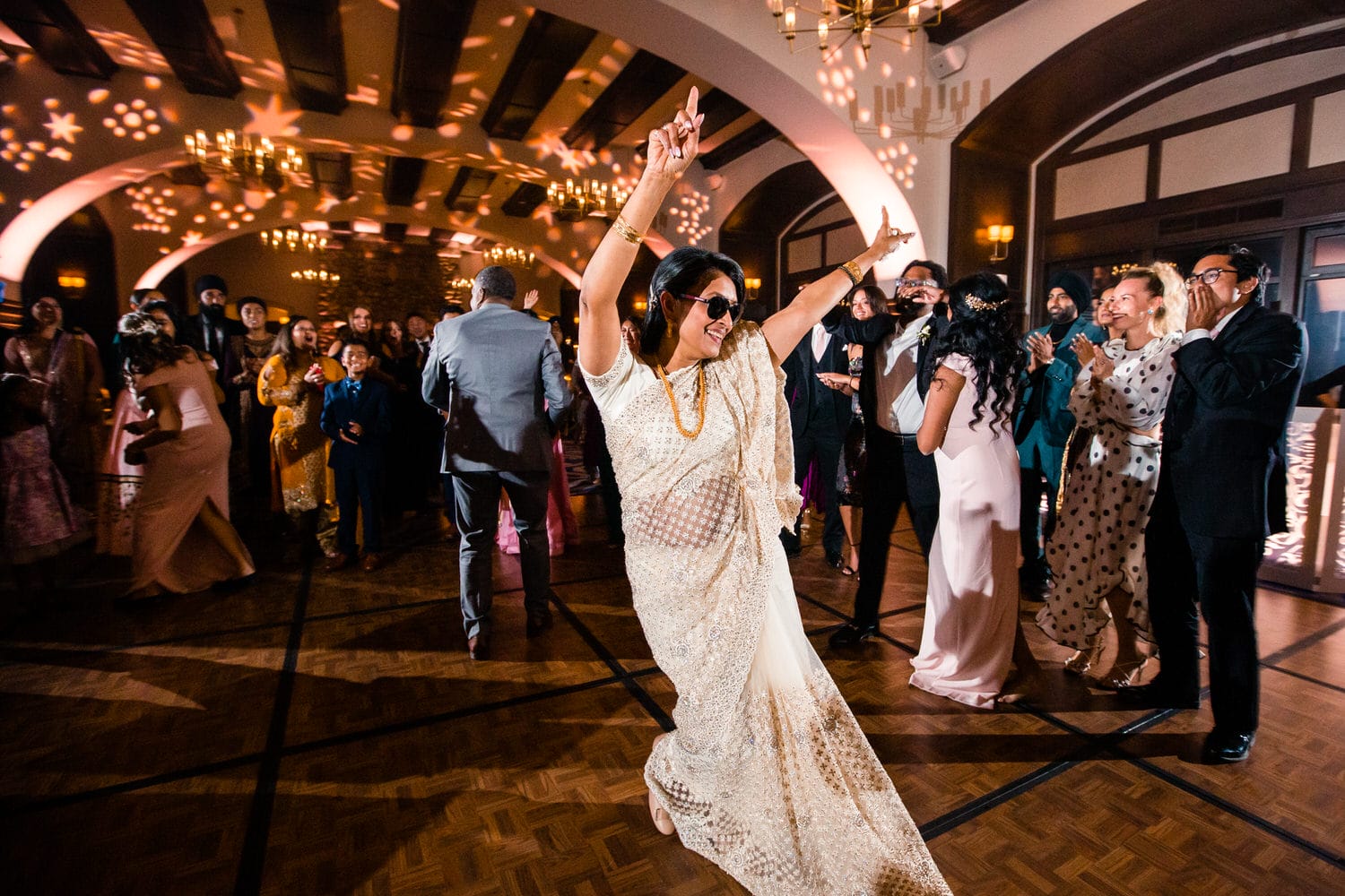 A joyful woman in a sequined outfit dances enthusiastically at a vibrant party, surrounded by guests celebrating with excitement.