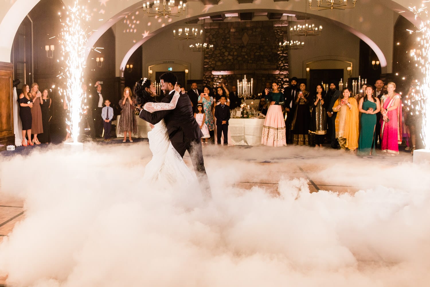 A couple shares a romantic dance surrounded by guests, with smoke and sparkler effects creating a magical atmosphere.