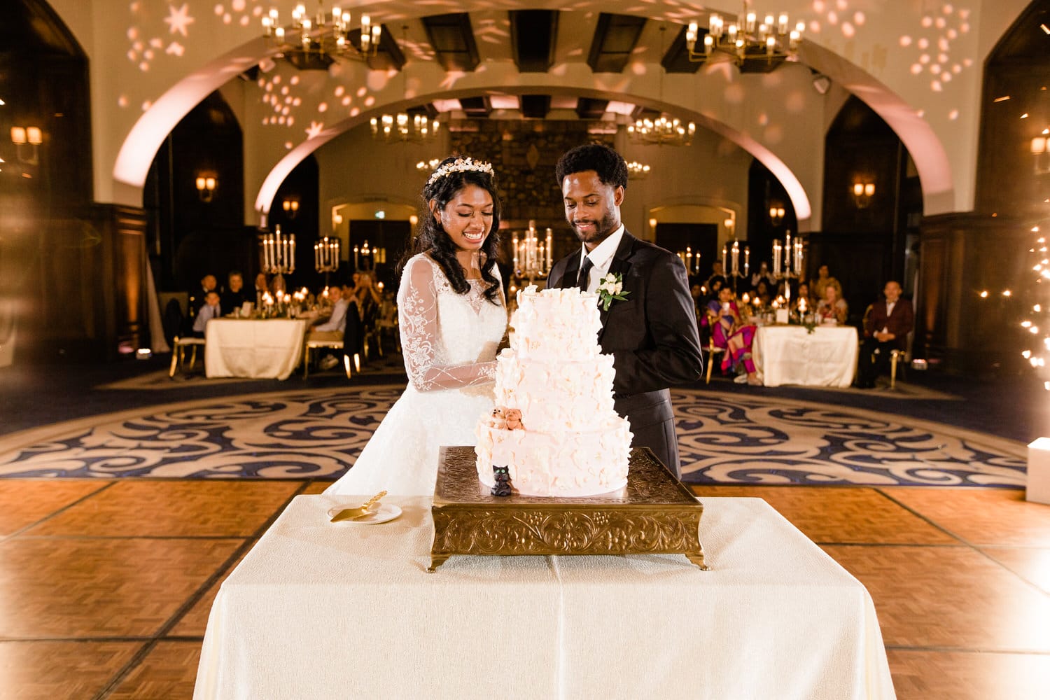 A joyful couple cuts their beautifully decorated wedding cake amidst an elegant reception setting, surrounded by family and friends.