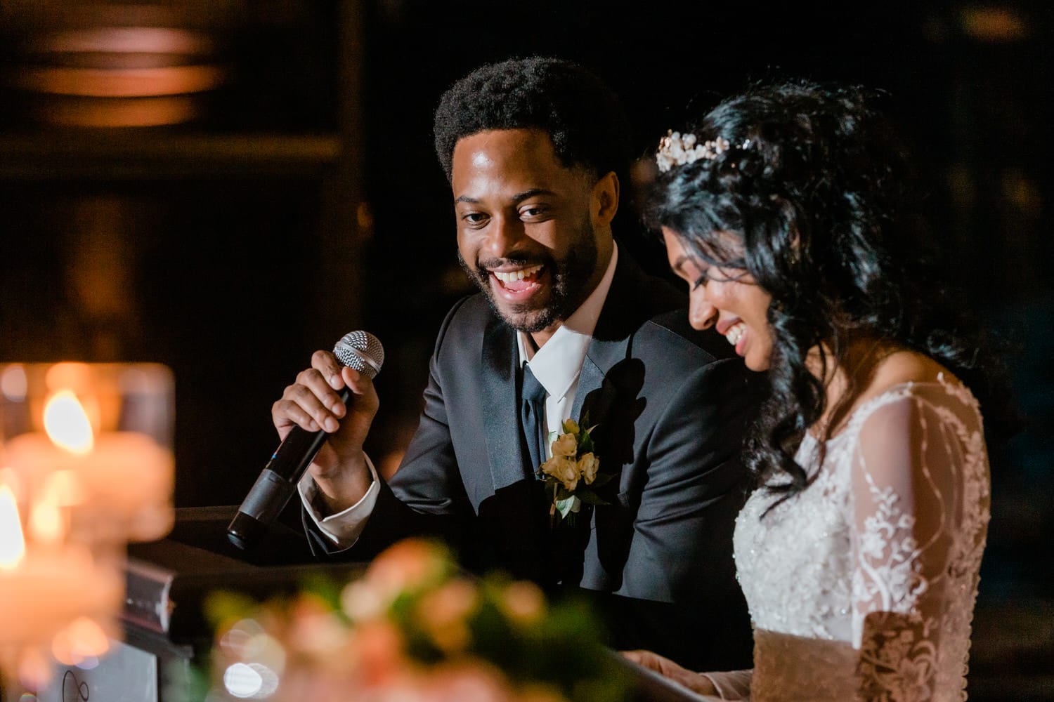 A groom smiles and speaks into a microphone while the bride, seated beside him, beams with happiness during their wedding celebration.