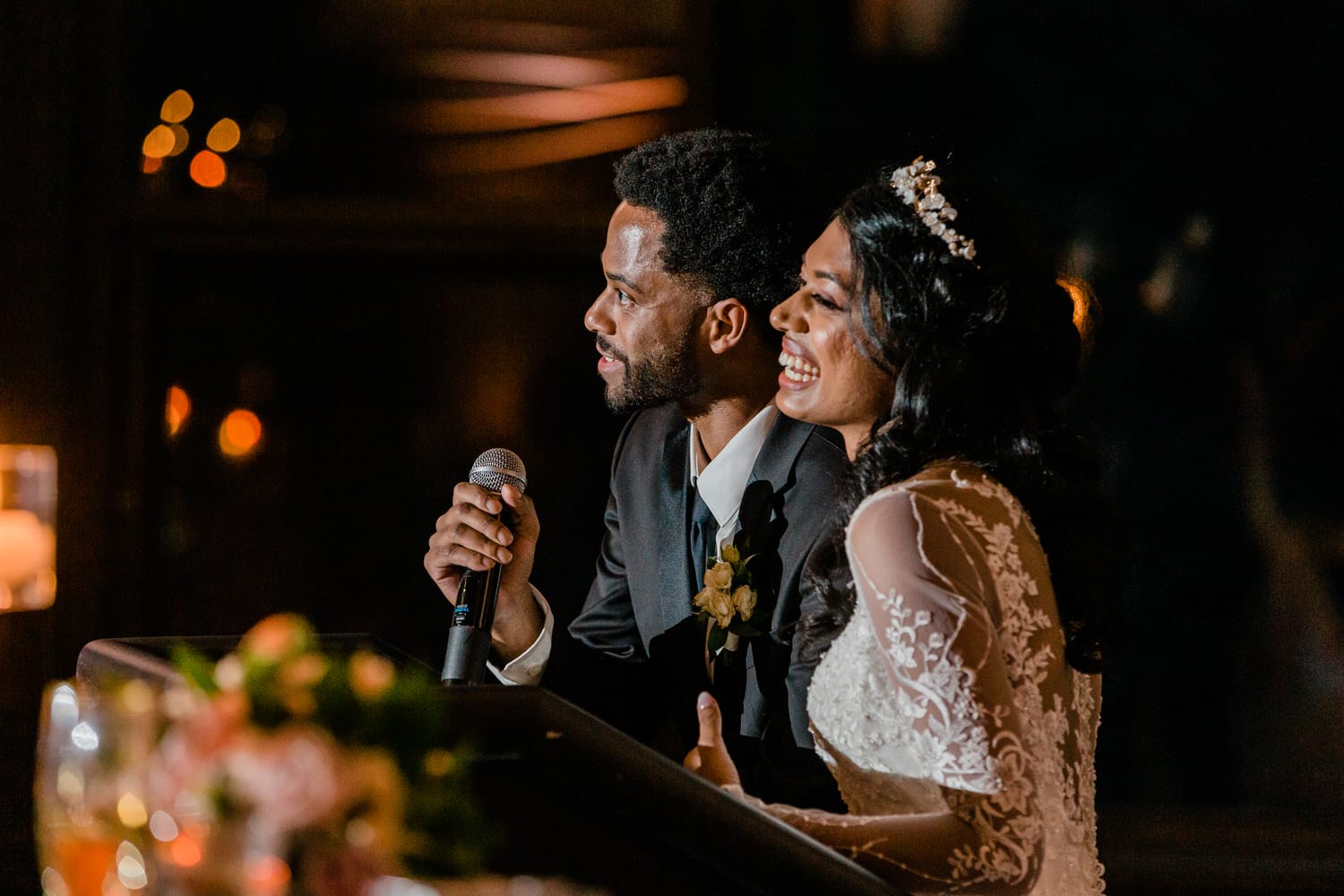 A couple joyfully sharing a moment while giving a toast during their wedding reception, with warm lighting and floral arrangements in the foreground.