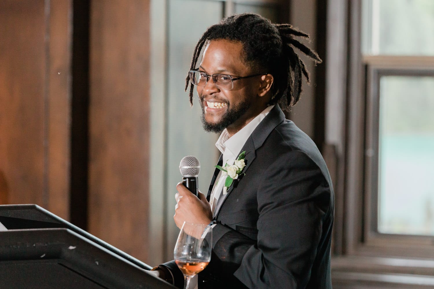 A man in a suit giving a speech with a microphone, smiling, and holding a glass of rosé wine in a warmly lit indoor setting.