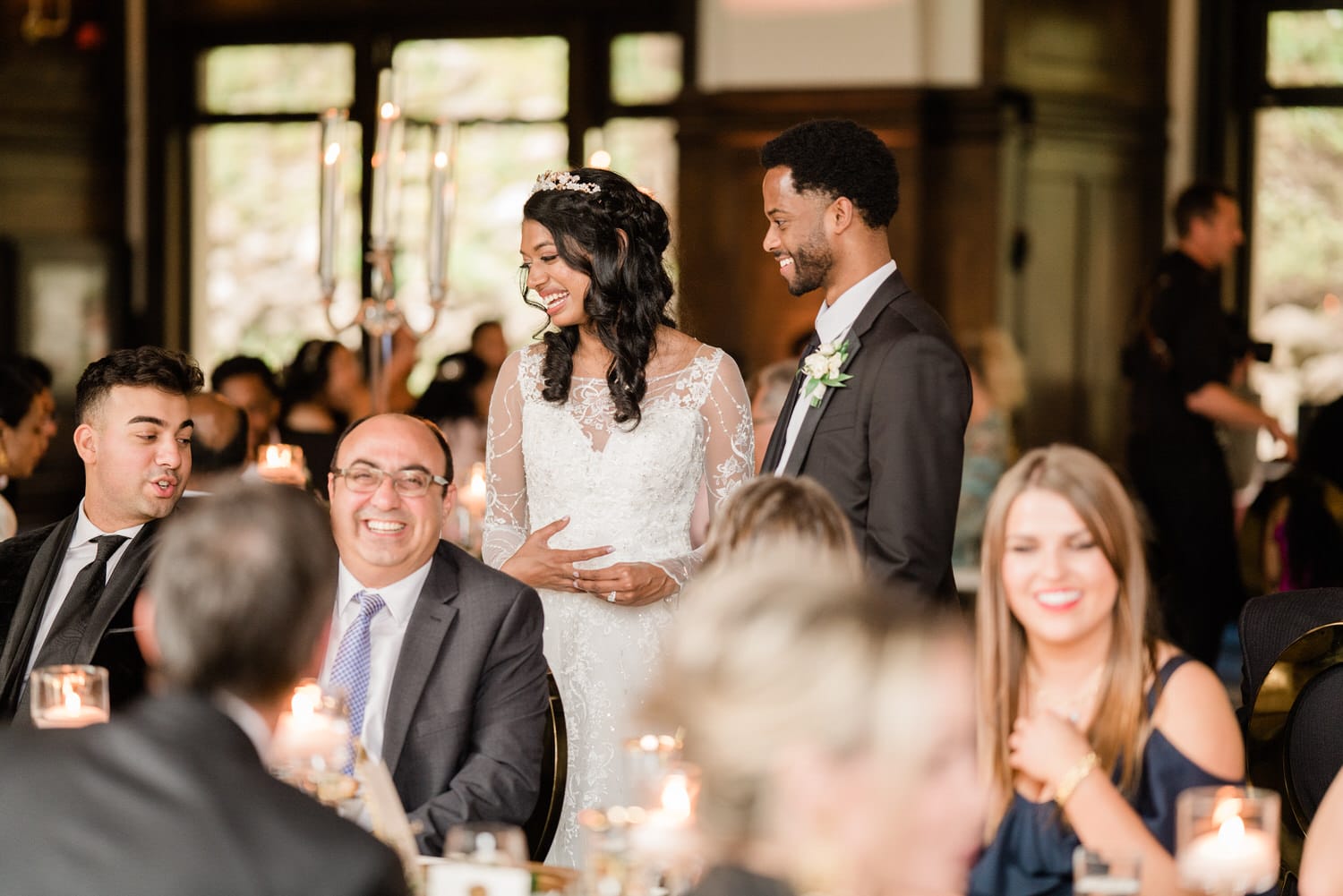 A smiling bride in a lace gown interacts with guests at a joyful wedding reception, surrounded by family and friends.