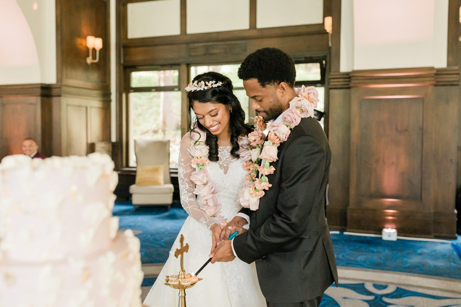 A couple in wedding attire smiles as they light a candle together, surrounded by floral leis, with a decorated cake in the foreground.