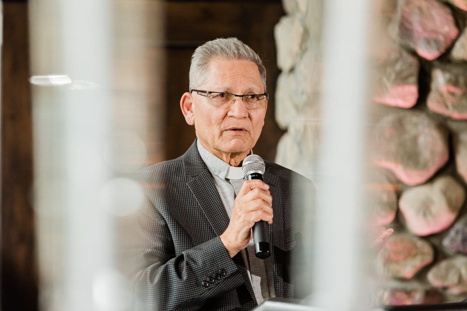 An older man in a suit holds a microphone while speaking, with a stone wall in the background.