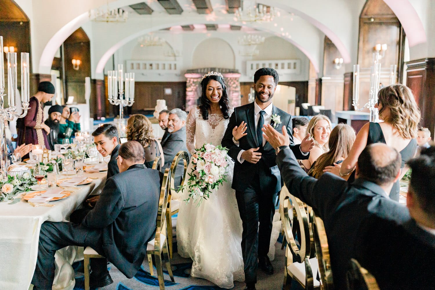 A newly married couple walks hand in hand through a festive reception, surrounded by smiling guests and elegant decor.