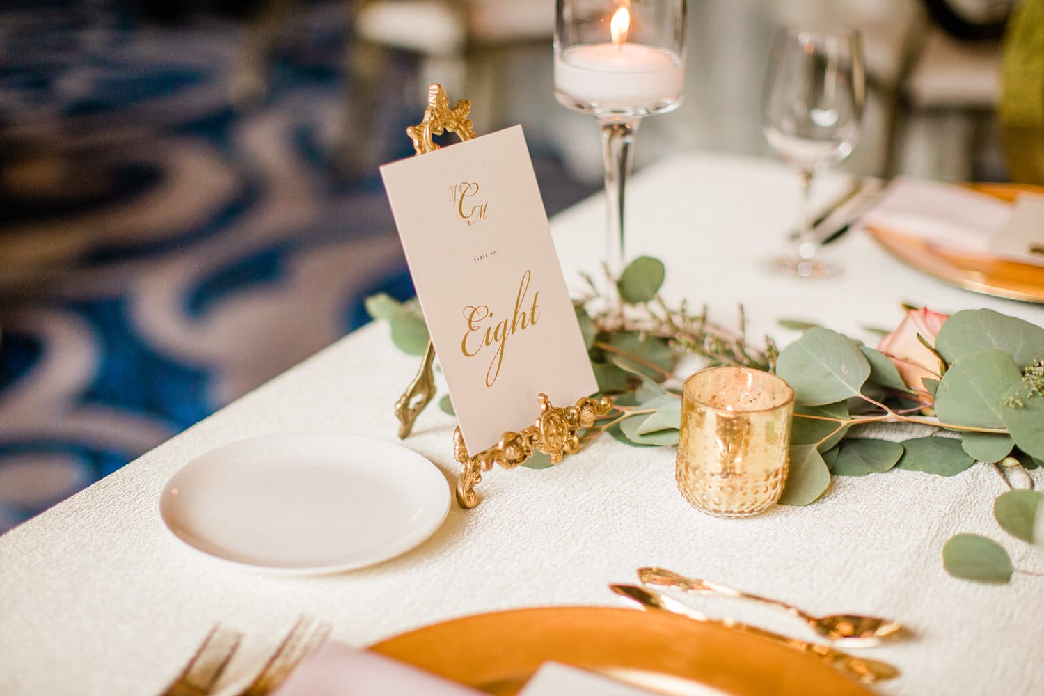 A beautifully arranged table featuring a gold-framed table number card reading "Eight," surrounded by greenery, a lit candle, and elegant dinnerware.