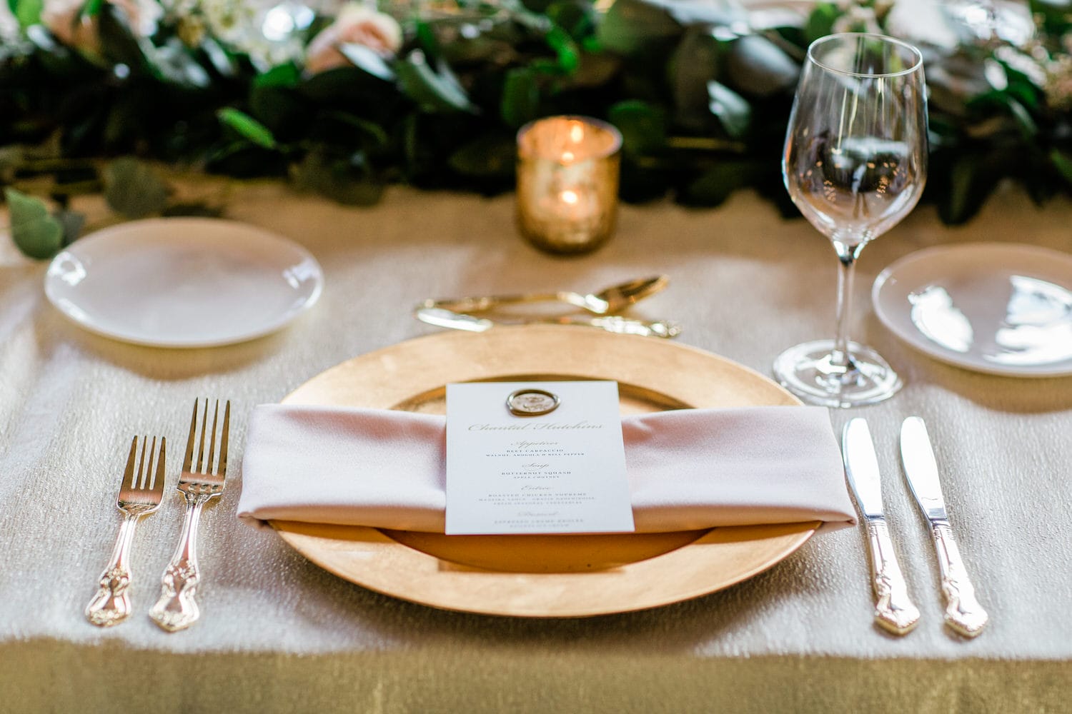 A beautifully arranged table featuring a gold charger plate, folded napkin, menu card, silver cutlery, and a wine glass, set against a backdrop of greenery and soft lighting.