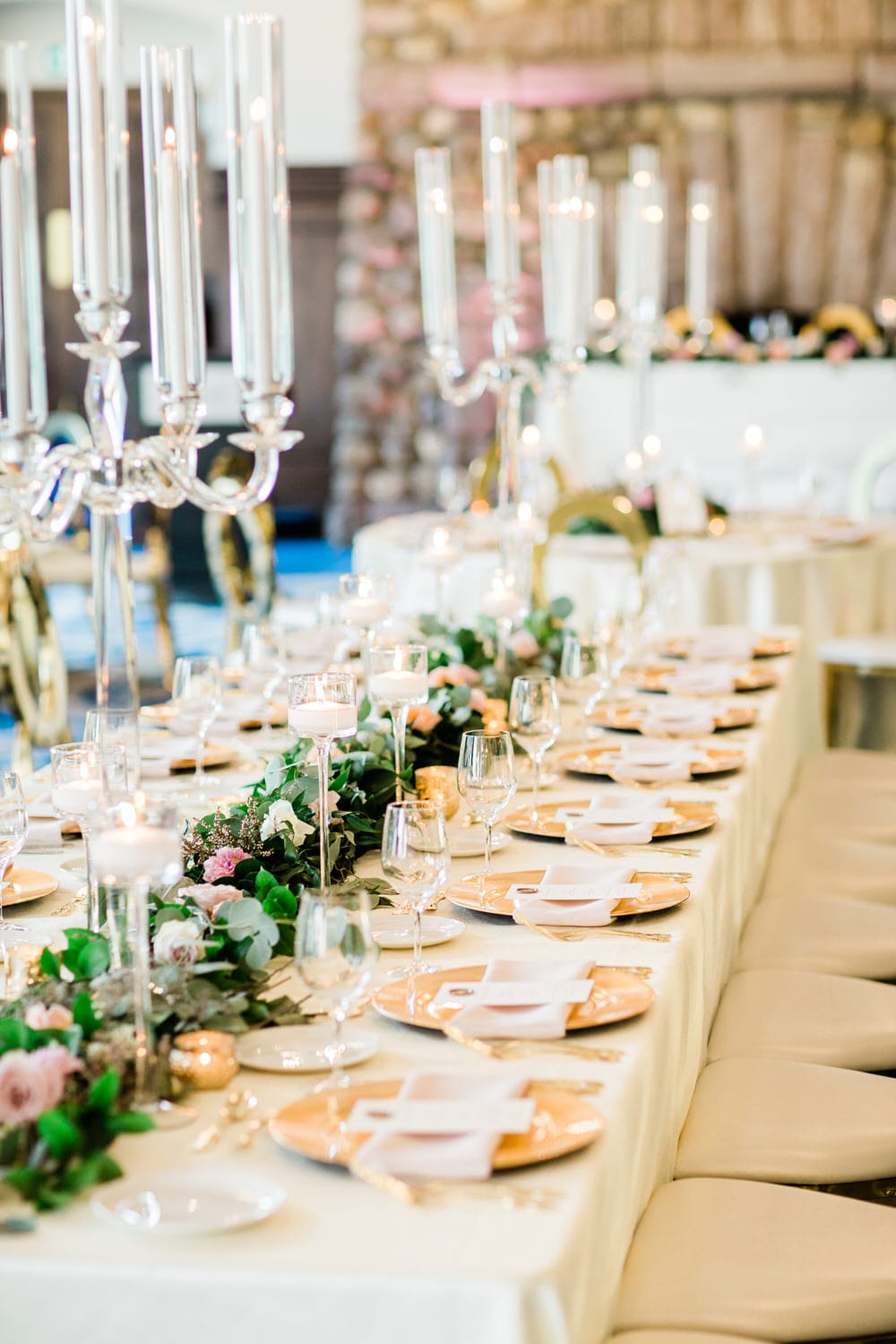 A beautifully arranged long dining table adorned with tall candlelit centerpieces, decorative plates, and floral accents, creating a sophisticated atmosphere for a wedding reception.