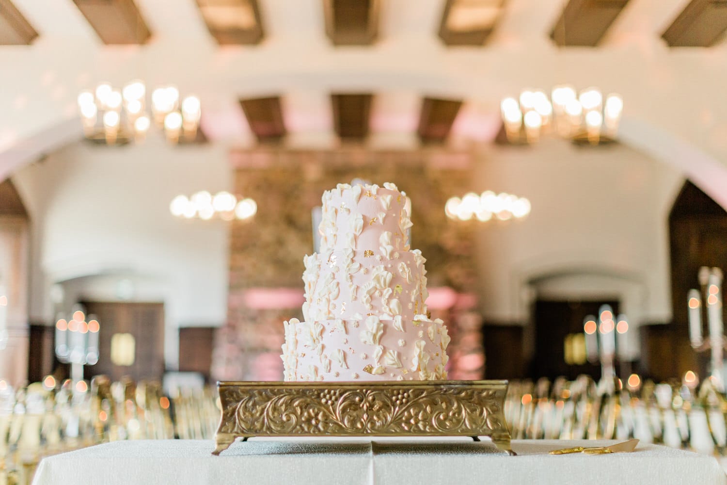 A beautifully decorated three-tiered pink wedding cake adorned with delicate floral details, presented on an ornate gold stand in a stylish reception venue.