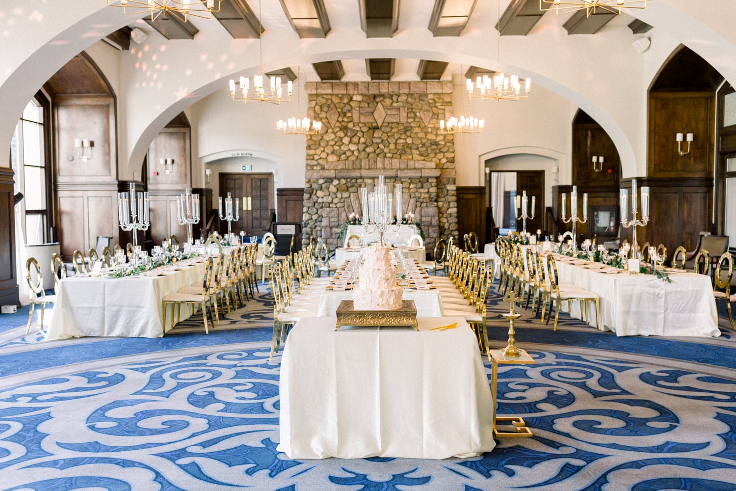 A beautifully arranged banquet hall featuring long tables with white linens, gold accents, and a stunning cake centerpiece, set against a backdrop of stone walls and chandelier lighting.