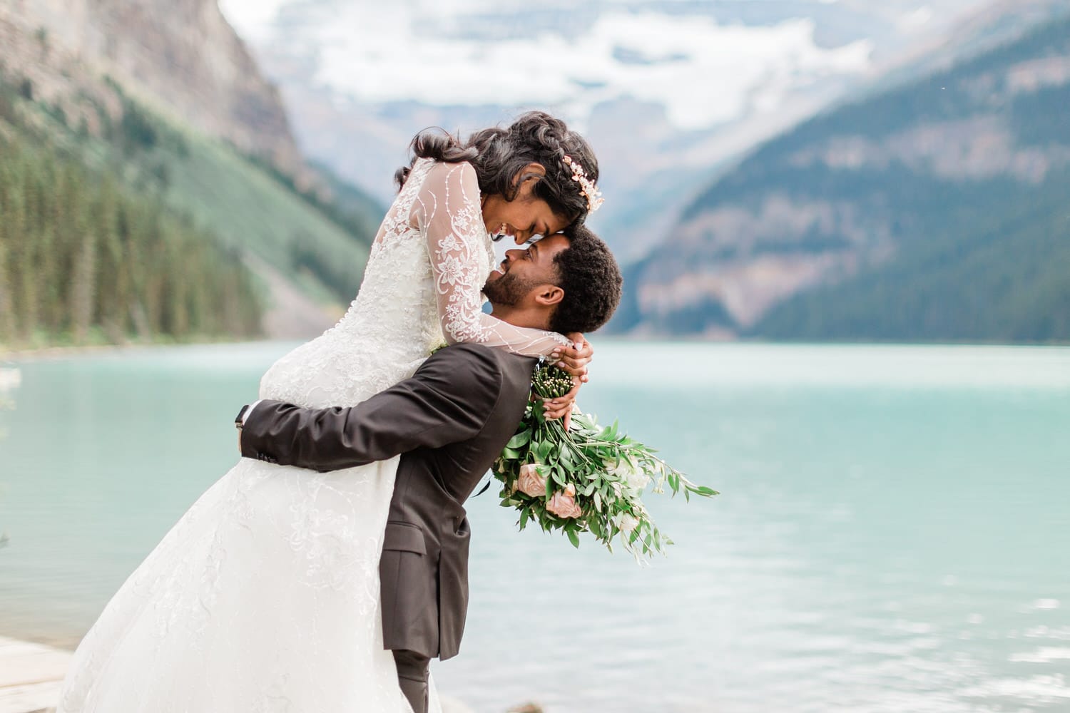 A couple shares a joyful moment by the lake, with the bride lifted in the groom's arms, surrounded by stunning natural scenery and vibrant greenery.