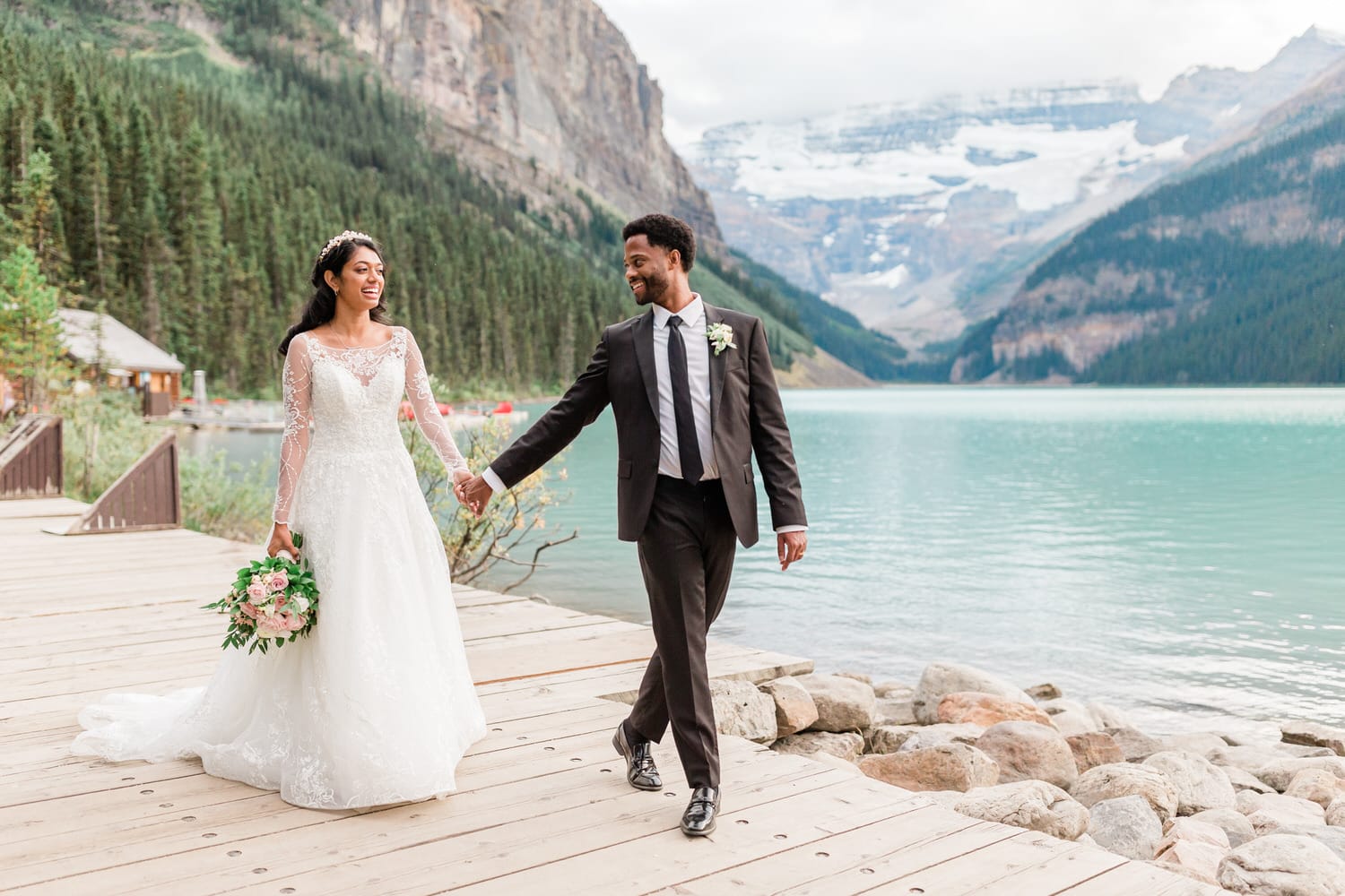 A joyful couple walks hand in hand along a wooden path by a serene lake, surrounded by mountains and evergreen trees.